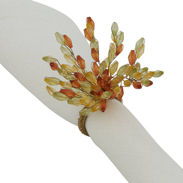A gold-toned napkin ring bracelet clustered translucent amber, orange, and pale yellow leaf-shaped beads, shown worn on a white fabric napkin.