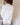 Model is turned away from the camera showing the back of the garment. She is wearing a white button up top and denim pants. The back of the shirt is white and you can see the seam down the middle and across the shoulders. Where the tag is sewn in, red thread is visible at the corners.
