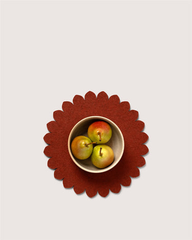 A top-down view of a scalloped dark red felt placemat with a small beige bowl containing three pears, set against a solid light gray background.