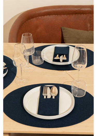A styled dining table setting featuring navy blue oval placemats, white plates, a navy napkin with silver cutlery, and clear drinking glasses, with a brown leather upholstered chair in the background.
