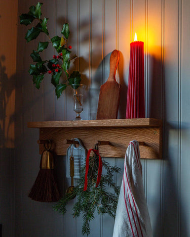 This image shows a shelf with hooks upon which sits a Greentree Petite Pillar candle in Red, lit. It sits amongst other wintery items.