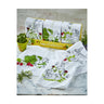 A set of white cotton dishtowels with green and red floral and bunny designs, displayed on a counter with some plants in the background.