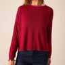 A woman wearing a long-sleeve knit sweater in deep burgundy red, paired with dark blue jeans, shown from the side against a plain beige background.