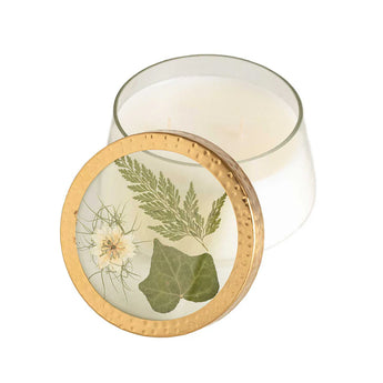 Clear glass container with off-white candle inside. Lid is presented in front of the candle. The Lid features a gold rim and clear glass center with pressed  botanicals in green hues.