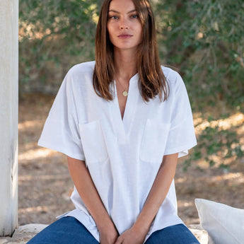 Woman with long brown hair seated wearing Mersea white pocket tee and blue jeans. Behind her are some trees.