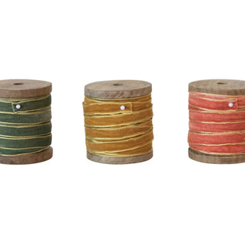 Three wooden spools with velvet ribbon in three different colors: green, gold and coral. Each has a metallic gold edge on the ribbon and they’re secured to the spool with a pin.