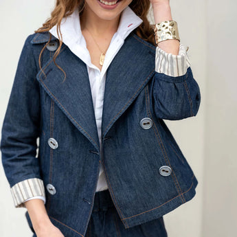 Model is wearing a standard blue denim colored peacoat, open, over a white shirt with her collar popped. She has the sleeves rolled up, revealing the striped lining of the coat. She has one hand raised, touching her hair, and she has a gold bracelet on that arm. The other arm is down at her side. She’s smiling and looking down.