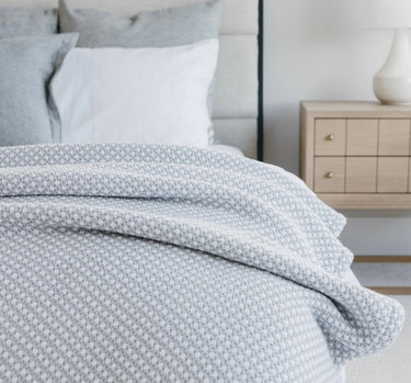 Image shows Faribault Mill’s Edgecomb blanket draped across a bed. The bed has several pillows on it. There is a nightstand with a lamp on it in the background.
