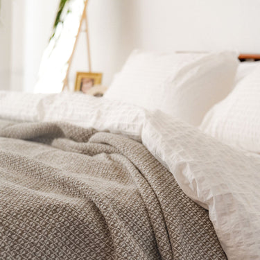 Image shows Faribault Mill’s Bar Harbor blanket draped across a bed. There is also a white comforter and white pillows on the bed. A nightstand with a photo frame is just out of focus in the background.