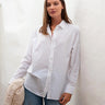 Brunette model is leaning up against a white wall. She is wearing a white button up top, light denim pants and is carrying an off-white cable-knit sweater.