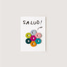A white greeting card with the word 'SAUD!' in black handwritten-style text and a cluster of colorful circular cutouts arranged like a flower, shown against a light warm gray background.