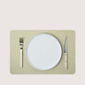 A minimalist place setting featuring a light sage green rectangular placemat, a white ceramic plate, a white-handled fork, and a white-handled knife, arranged symmetrically on a solid light beige background.