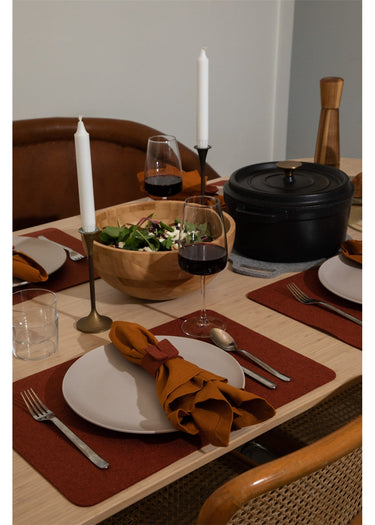 A styled dining table setting with white plates, silver cutlery, brown placemats, a folded rust-orange napkin, a wooden salad bowl, wine glasses, and a black Dutch oven on a light wood table.