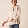 A woman modeling a light beige long-sleeve top with a split V-neckline and buttoned cuffs, paired with blue jeans, against a solid light beige background.