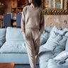 A woman wearing a matching beige knit sweater and jogger pants set stands in a living room in front of a light blue sofa.
