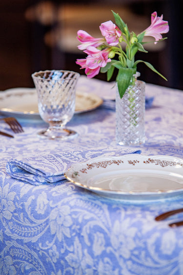 A styled table setting featuring a light blue and white damask patterned tablecloth, white plates with gold trim, a blue napkin, and a small clear glass vase holding pink alstroemeria flowers.