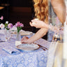 A close-up lifestyle shot of a person seated at a table set with a light blue and white damask patterned tablecloth, silver plates, glassware, and small pink flowers.