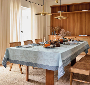 Manor Lane Blue Collection Tablecloth on table. Main area of tablecloth is light blue with a floral damask pattern. The border features a floral/geometric design in a lighter and darker blue. Shown on a set table