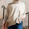 A woman wearing a beige ribbed turtleneck sweater and blue jeans, holding a brown leather clutch in an outdoor setting.