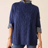 A woman wearing a navy blue cable-knit mock-neck sweater with striped knit cuffs, layered over a matching striped mock-neck shirt, paired with blue jeans against a plain beige background.