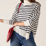 A woman wearing a cream and navy blue horizontally striped knit turtleneck sweater, paired with blue jeans and holding a red shoulder bag, against a plain off-white background.